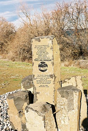 [A tall thin stone stands in the middle of shorter stones connected by a ring of metal wire. There are pebbles around the marker which sits in the middle of a grassy section near the river.]